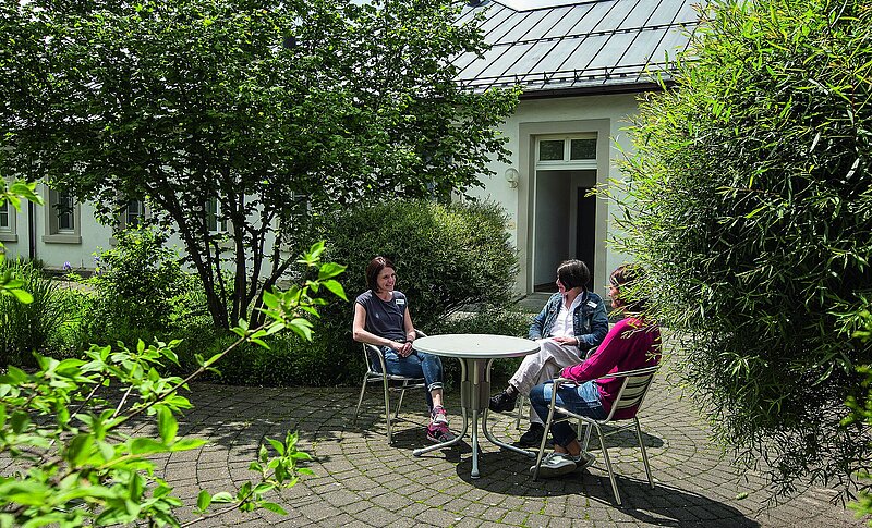 Vor einem Gebäude sitzen drei Frauen im Garten an einem runden Tisch. Sie unterhalten sich.