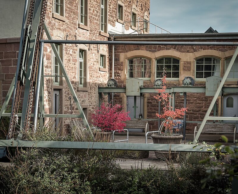 Historisches Gebäude der Tagesklinik, im Vordergrund sind Stahlelemente sichtbar. Der Hof ist bunt bepflanzt.
