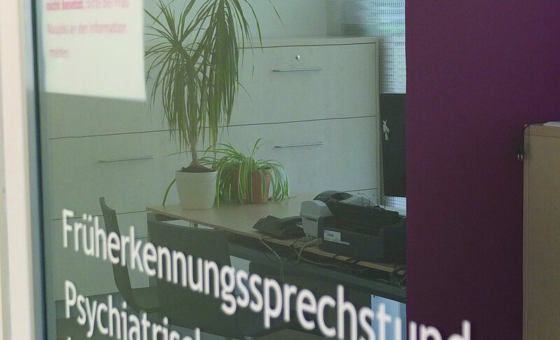Durch ein großes Fenster blickt man auf einen Schreibtisch mit Telefon und zwei Pflanzen. Das Fenster ist beschriftet mit Früherkennungssprechstunde und Psychiatrische Institutsambulanz. 