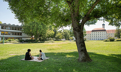 Zwei Menschen sitzen auf einer Decke neben einem Baum in der blühenden Parkanlage. Im Hintergrund sind verschiedene Klinikgebäude zu sehen.