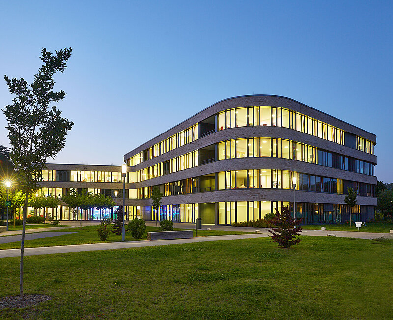 Im Dämmerungslicht strahlt das hell beleuchtete Klinikgebäude. Umgeben von grüner Wiese und jungen Bäumen.