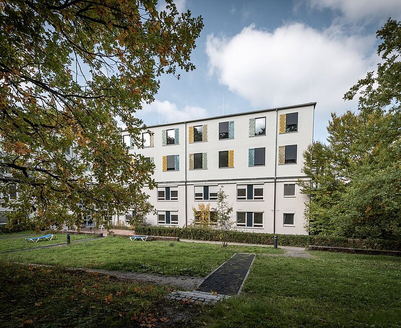 Ansicht auf ein modernes vierstöckiges Behandlungszentrum mit gelben, blauen und grünen Fensterläden, umrandet von Bäumen. Im Garten stehen einige Liegestühle.