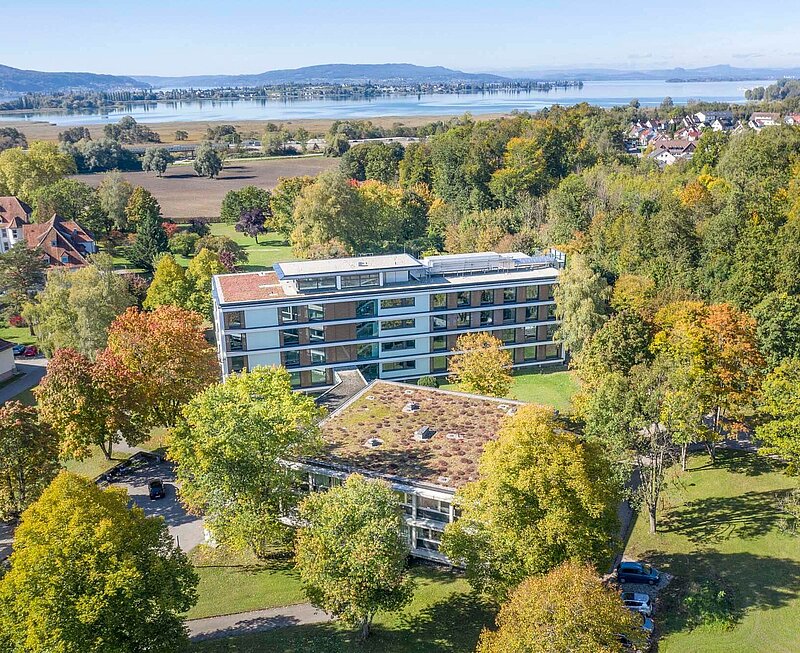 Zwei mehrstöckige Klinikgebäude sind mit einem Durchgang verbunden. Das Gebäude ist Teil eines größeren Geländes. Im Hintergrund ist der Bodensee zu sehen.