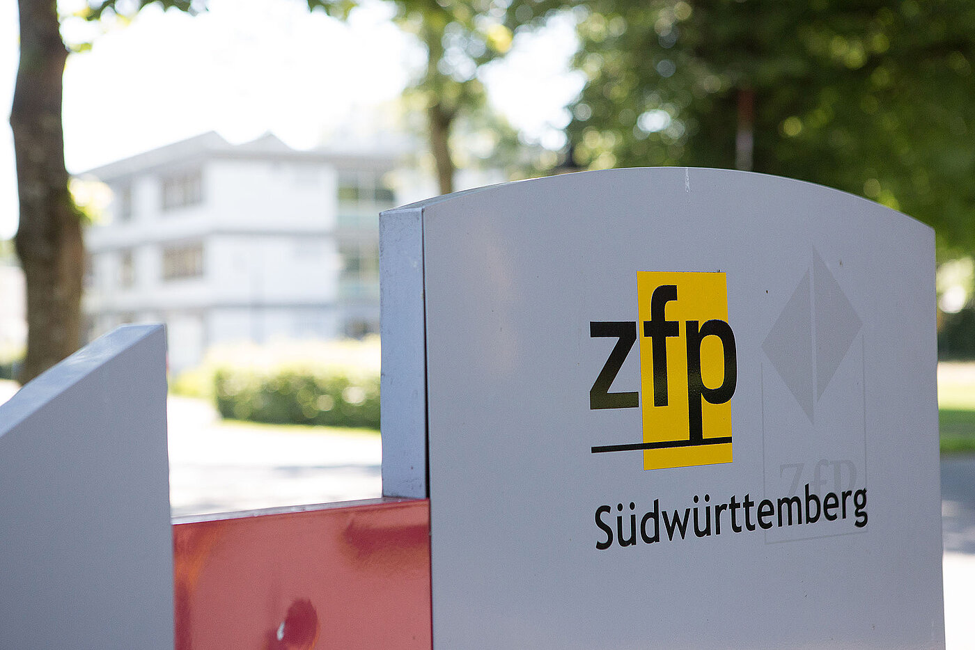 Metalschild mit dem Logo zfp. Das bedeutet Zentren für Psychiatrie. Im Hintergrund sind Klinik und Bäume sichtbar.