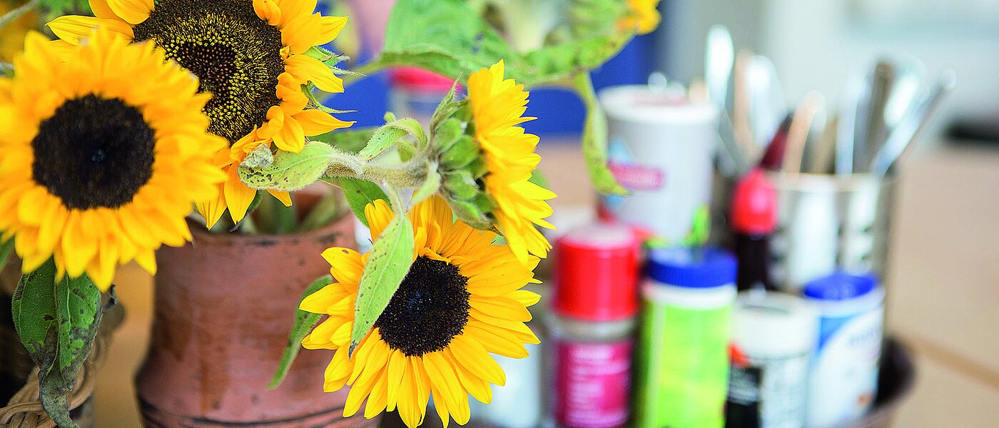 Vase mit Sonnenblumen steht auf einem Tisch. Auf einem Tablett stehen verschiedene Gewürze in farbigen Verpackungen.