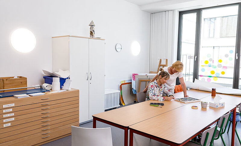 Im Raum befinden sich mehrere Tische, ein weißer Schrank und ein großer Schubladenschrank mit Zeichenutensilien. Am Tisch sitzt eine Frau. Sie malt auf ein Blatt. Hinter ihr steht eine Therapeutin. 