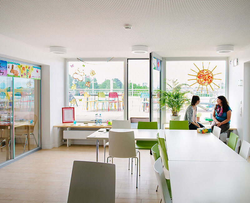 In einem großen hellen Raum sitzen zwei Frauen auf der Fensterbank. Im Raum stehen Tische und Stühle, die Fenster sind mit Bildern und Fensterbildern beklebt. Vom Raum geht eine Tür raus zu einer großen Terrasse.