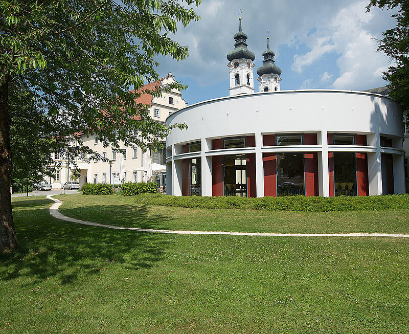 Ein Gebäude mit runder Fassade ist umgeben von einer Grünanlage. Im Hintergrund sind Teil der Klosteranlage zu sehen.