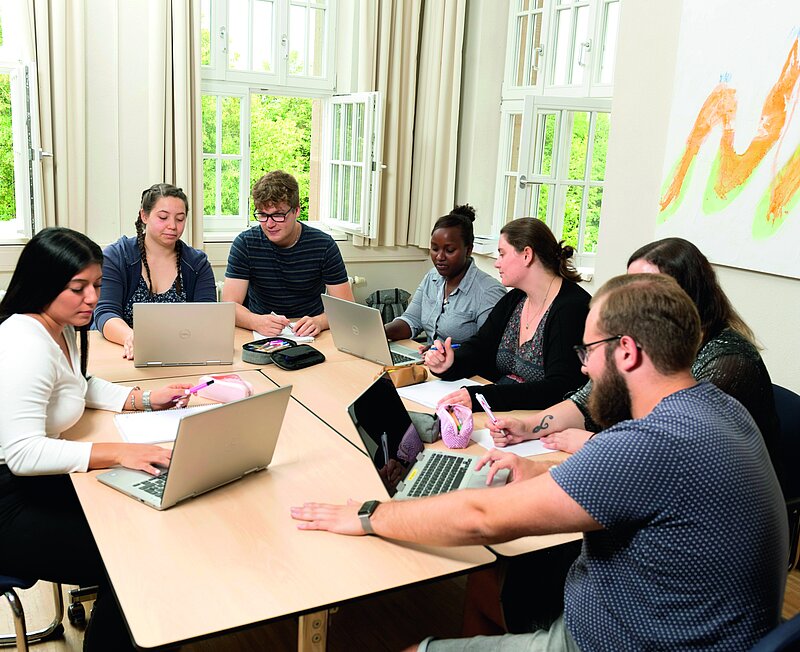 Fünf Frauen und zwei Männer, alle noch jünger, sitzen in Kleingruppen am Tisch und diskutieren miteinander. Dazu nutzen sie auch vier Laptops.