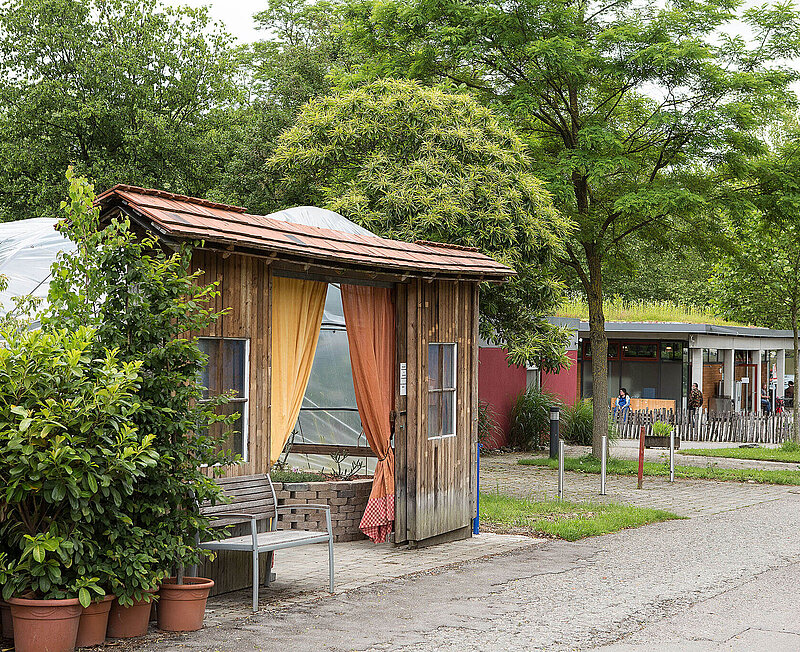 Der Eingang zu den Gebäuden der Werkstätten ist umsäumt von vielen grünen Topfpflanzen. Im Hintergrund sind Menschen und ein Parkplatz zu sehen.