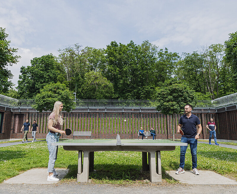 Durch einen hohen Zaun geschützer Innenhof. Hier spielen zwei Personen Tischtennis. Weitere Personen spazieren auf dem Gehweg. Zwei Personen sitzen auf einer Gartenbank und unterhalten sich.