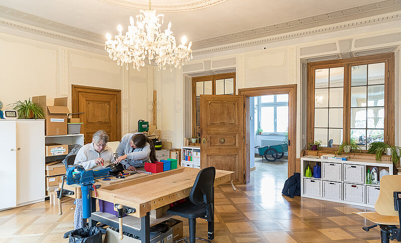 In einem historischen Werkstattraum mit großem Kronleuchter an der Decke steht eine Werkbank. Hier sitzt eine Frau und schraubt an einem defekten Gerät. Eine Therapeutin steht unterstützend zur Seite.