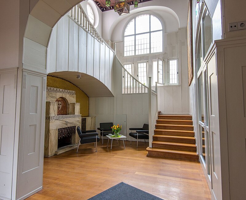 Historischer Raum mit einem Kamim und Sitzecke. Von hier führt eine hölzerne Treppe in das nächste Stockwerk.