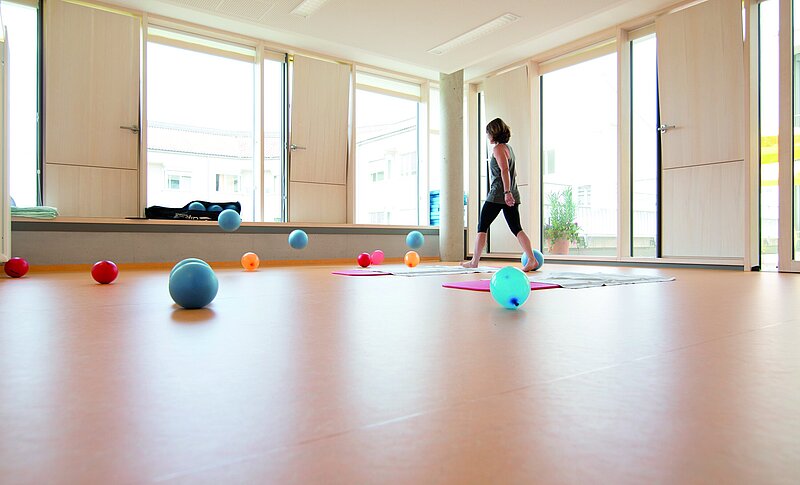 Im hellen Gymnastikraum liegen und fliegen bunte Bälle und Luftballons. Eine Frau in Sportkleidung bewegt sich über zwei Gymnastikmatten..