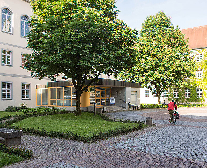 Ein Fahrradfahrer fährt über Kopfsteinpflaster zum Eingang des Klostergebäudes. Um den Eingang herum sind eine kleine grüne Fläche und Bäume.