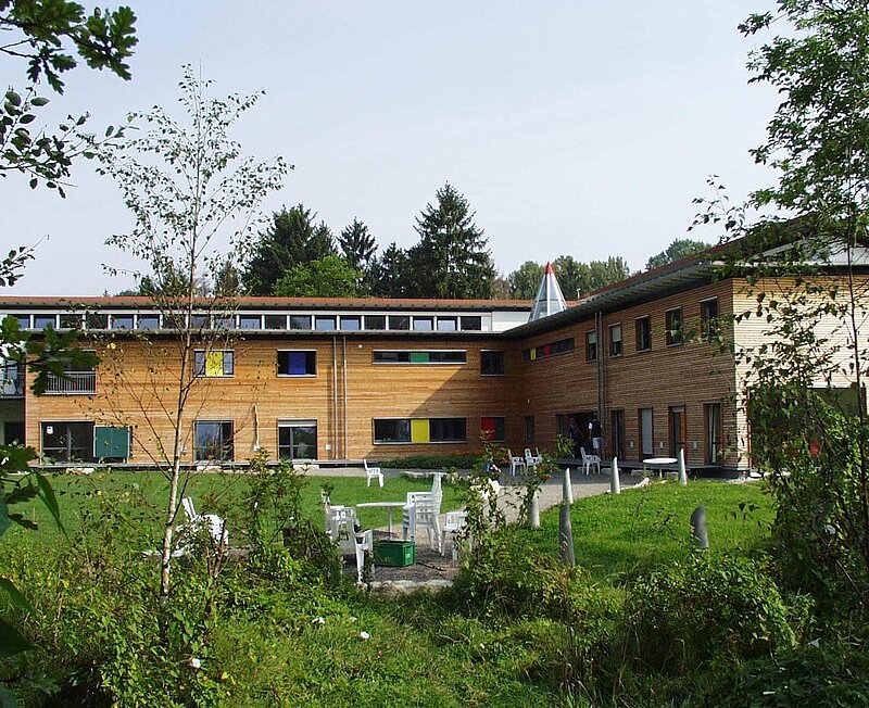 Im Hintergrund steht ein Gebäude mit Holzfassade. Im davor liegenden Garten gibt es verschiedene Sitzbereiche.