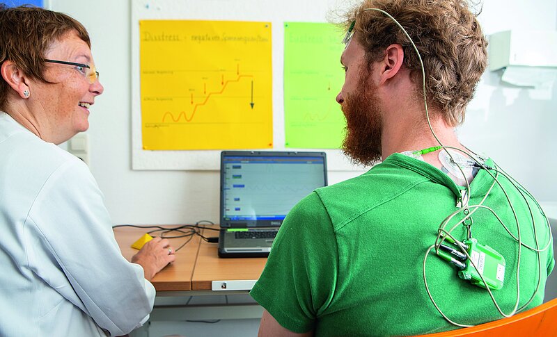 Eine Ärztin und ein Patient sitzen am Tisch, vor einem Computer. Der Mann ist am Kopf und Nacken mit Elektroden versehen. Die beiden unterhalten sich.
