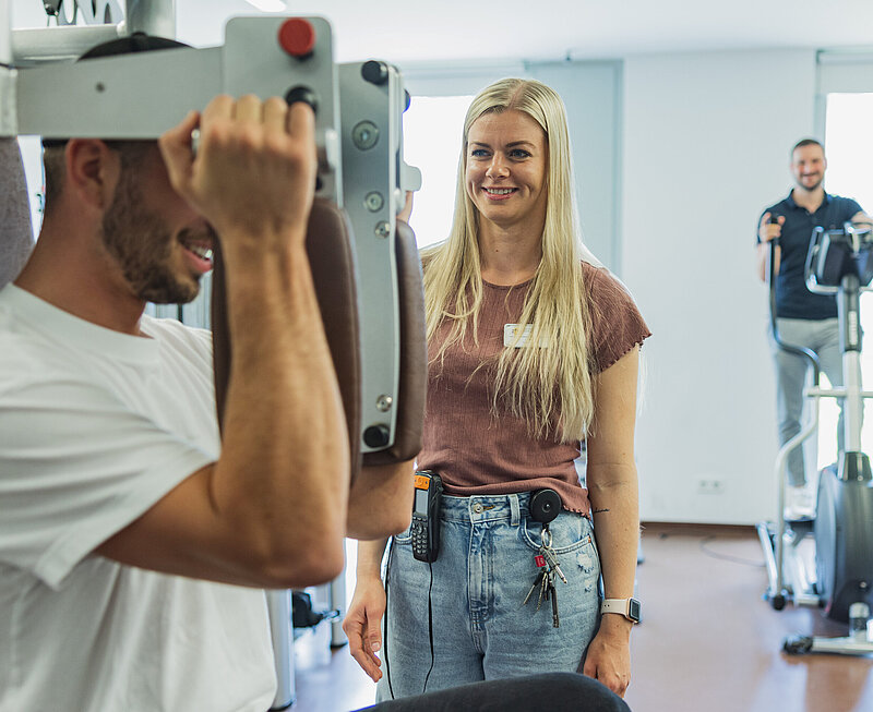 Zwei Männer trainieren im Fitnessraum. Eine Therapeutin steht neben dem vorderen Mann, der an einem Gerät für die Schultermuskulatur trainiert.