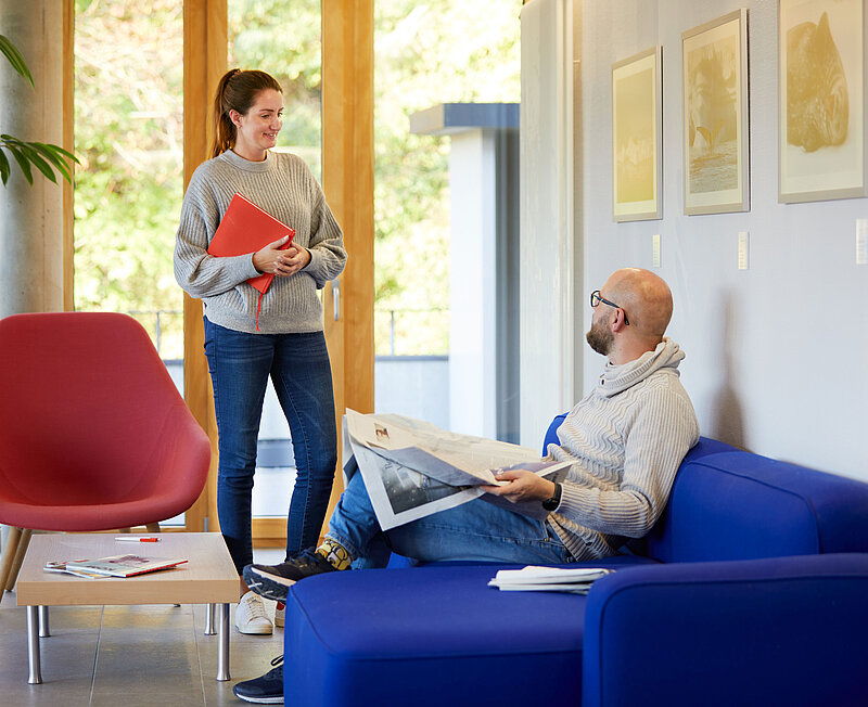 Ein Mann sitzt auf einem blauen Sofa mit der Zeitung auf dem Schoß. Er unterhält sich mit einer Frau, die neben ihm steht..