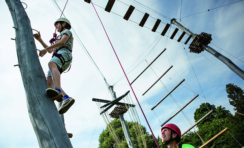 Ein junge klettert gut gesichert an einem Kletterpfahl hoch. Unten steht ein Mann und sichert den Jungen. Es handelt sich um einen Kletterpark mit mehreren Klettermöglichkeiten.
