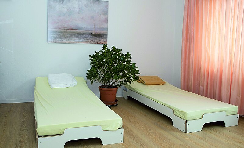 Das ist ein Patientenzimmer mit zwei Betten. Zwischen den Betten steht eine große Grünpflanze. An der Wand dahinter hängt ein Bild.