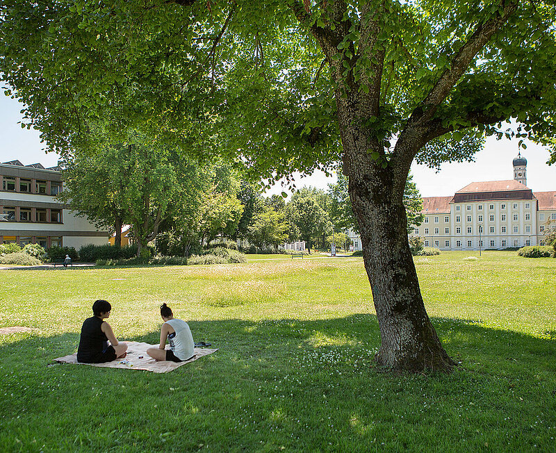 Zwei Menschen sitzen auf einer Decke neben einem Baum in der blühenden Parkanlage. Im Hintergrund sind verschiedene Klinikgebäude zu sehen.