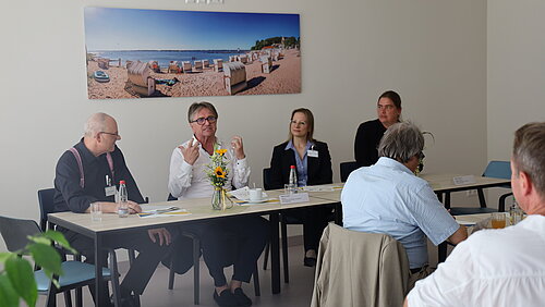 Der baden-württembergische Sozial- und Gesundheitsminister Manne Lucha MdL mit Andreas Breitmayer, Kaufmännischer Direktor und stv. Geschäftsführer des Klinikums am Weissenhof und der Dualen Leitung der Klinik für Allgemeine Psychiatrie und Psychotherapie im Klinikum am Weissenhof. Die vier Personen sitzen gemeinsam am Tisch. Im Hintergrund hängt ein Bild, vor ihnen sitzen Menschen die zuhören.