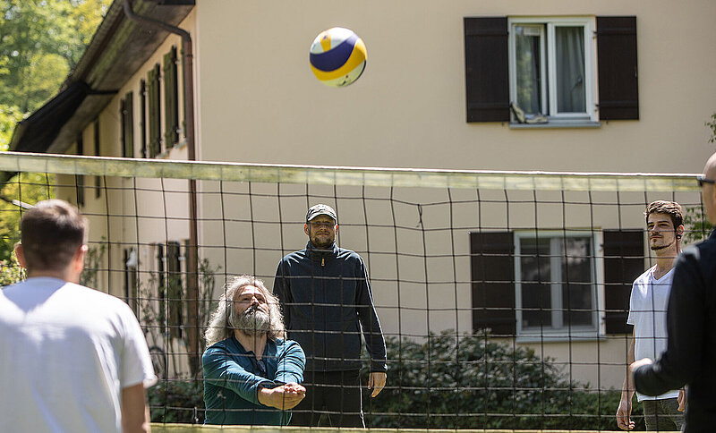 Eine Gruppe von Männern spielt draußen Volleyball. Im Hintergrund ist ein Gebäude zu sehen.
