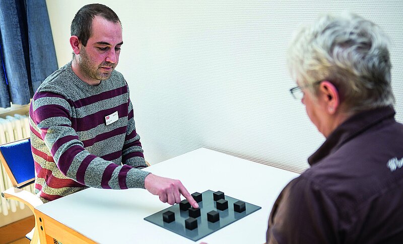 Links sitzt ein Mann, ihm gegenüber eine Frau an einem Tisch. In der Mitte befindet sich ein Brett mit schwarzen Würfeln. Der Mann macht einen sogenannten Block-Tapping-Test.