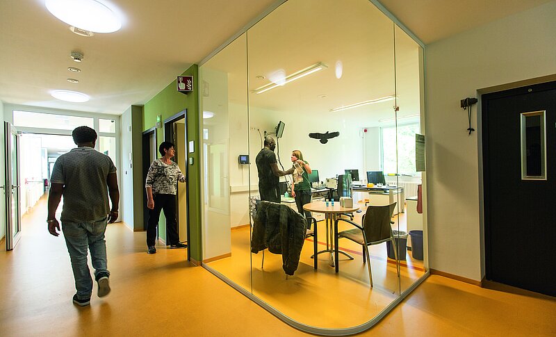Das ist eine Station der Forensischen Abteilung. Ein Mann bekommt im Mitarbeitendenzimmer ein Glas Wasser, eine ältere Frau wartet an der Tür. Auf dem Flur läuft ein Mann.