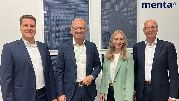 Drei Herren mit weißen Hemden und dunklen Jacketts und eine Frau im hellgrünen Blazer stehen nebeneinander. 