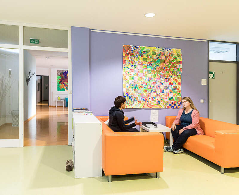 Zwei Frauen sitzen sich auf orangefarbenen Sofas gegenüber und unterhalten sich. An der Wand hängt Kunst.