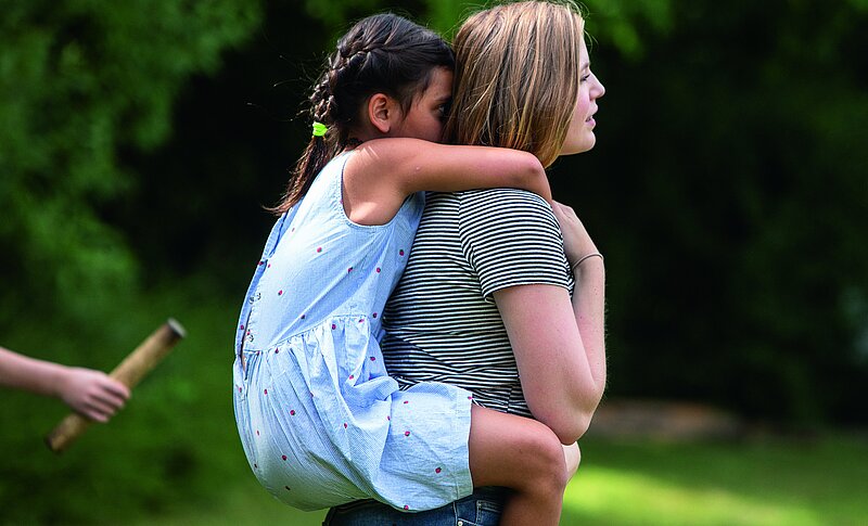Eine Frau trägt ein Kind Huckepack. Sie befinden sich draußen, in einem grünen Park.