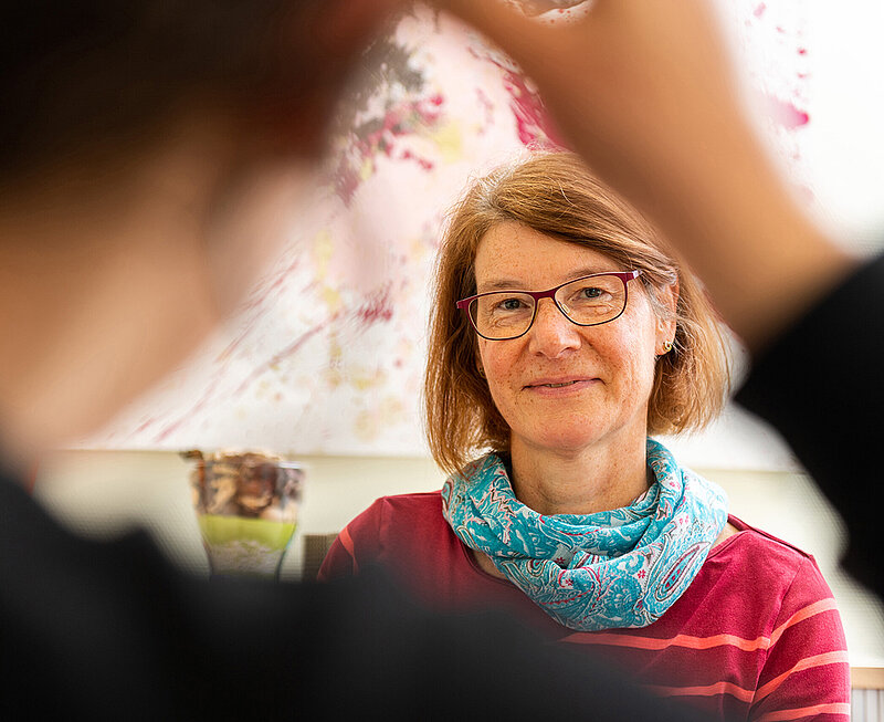 Eine Frau mit roter Brille sitzt am Tisch. Ihr gegenüber sitzt eine weitere Person.